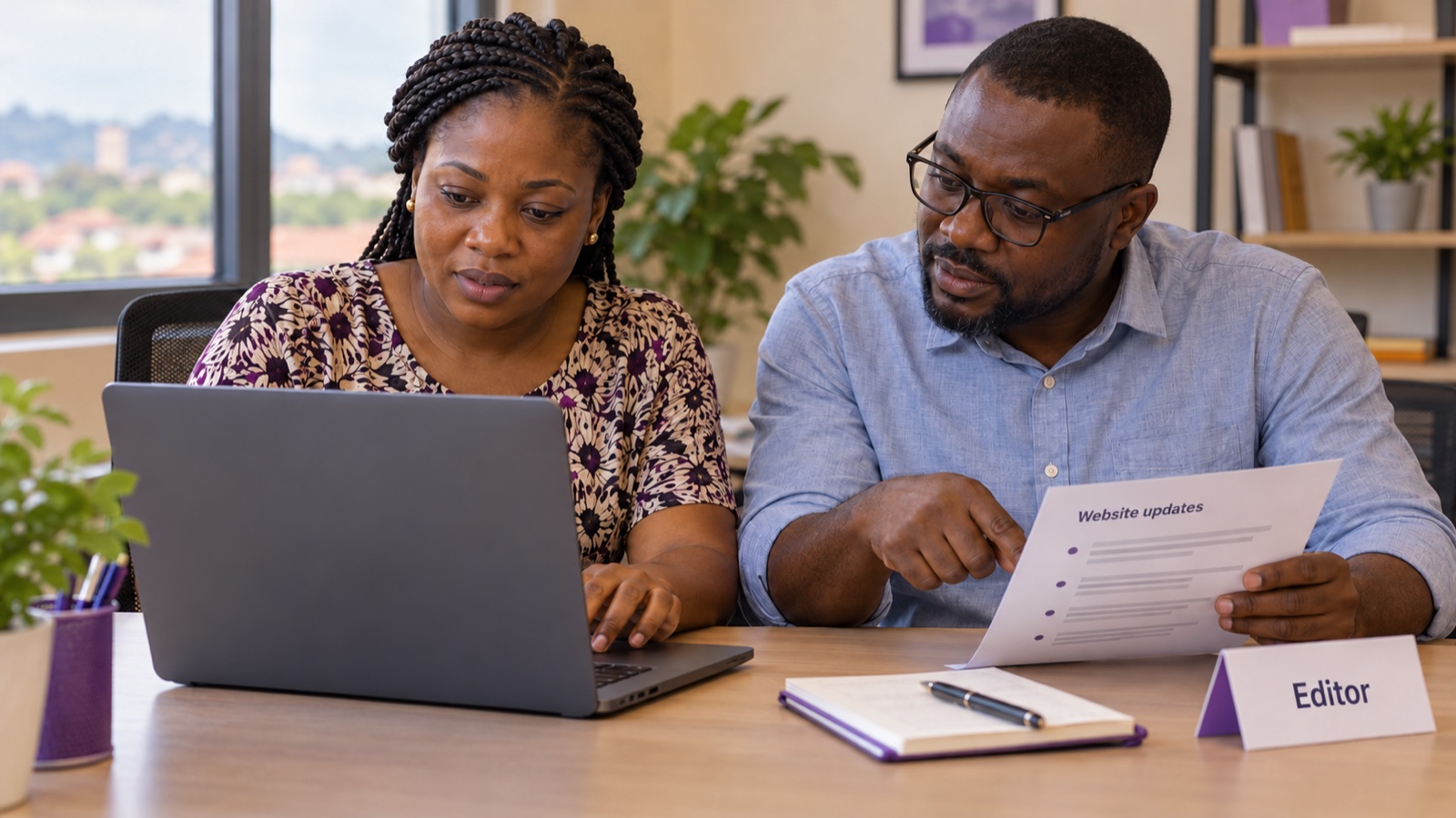 Two Ghanaian colleagues reviewing website update notes at a laptop beside an Editor role card.